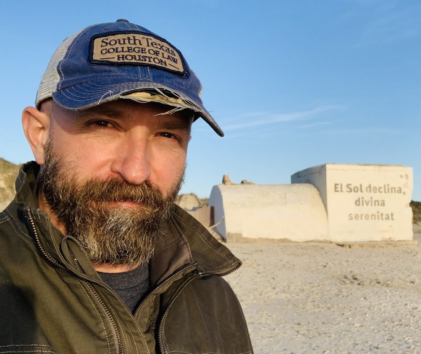 Russ in a South Texas College of Law cap beside a wall reading El Sol declina, divina serenitat